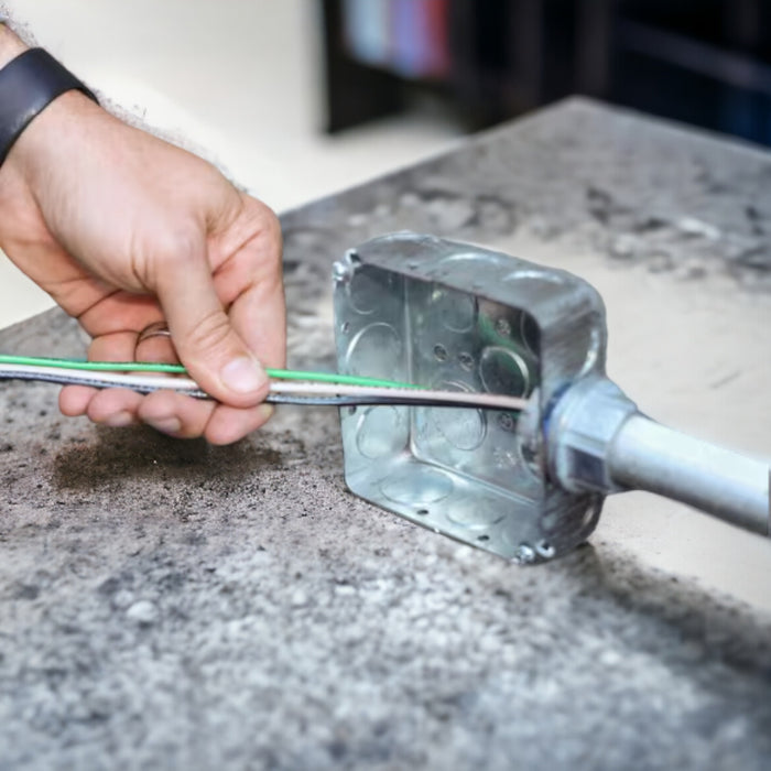A hand skillfully inserts Southwire 12 AWG WHT 500ft electrical wire into a metal junction box on a workbench, seamlessly connecting it to a metal pipe. The background is tastefully blurred, emphasizing the precise wiring task at hand.