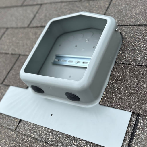 A close-up of a gray, empty, rectangular mounting bracket installed on a shingle roof for the SolaDeck 0799-2G JB-1.XL Junction Box. Manufactured by Soladeck, this bracket features side holes and includes a metallic component inside, possibly designed for PV roof-mount combiner equipment. It is securely supported by a flat white base on the roof.