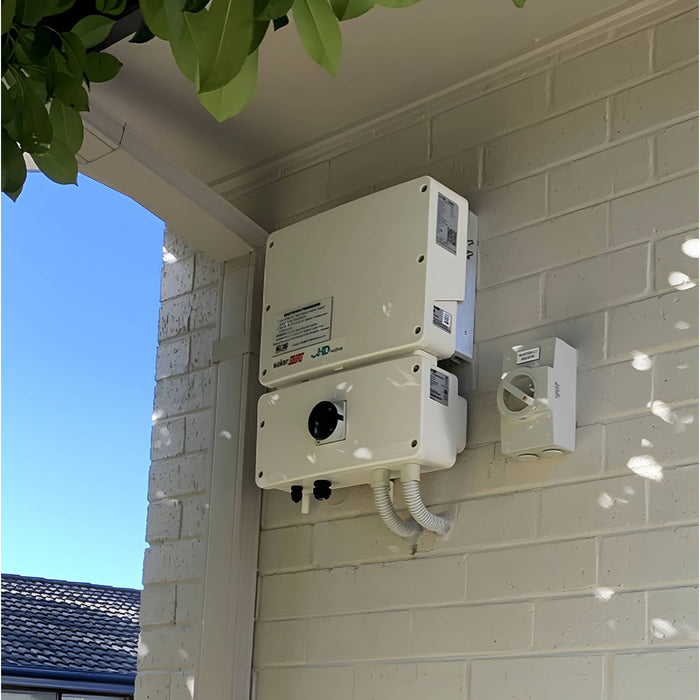 A white SolarEdge HD Wave 10kW Inverter SE10000H-US000BEI4, by Solaredge, is installed on a light brick wall outside, sheltered under an overhanging roof. A clear blue sky and tree leaves are visible in the background. Conduit pipes, optimized for efficient power flow, extend from the bottom of the inverter.