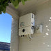 Outside a building, a white Solartek SolarEdge HD Wave Inverter 5kW and potential power optimizers are mounted on a brick wall. Above, green-leaved branches extend under a clear blue sky.