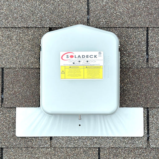 A SolaDeck 0799-2G JB-1.XL Junction Box by Soladeck, ETL listed for safety, is mounted on a shingled roof. The junction box features warning labels and has a sleek gray metal finish.