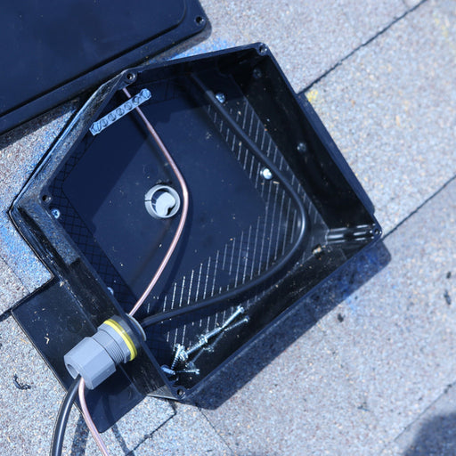 An EZ Solar JB-1.XL Junction Box in black with visible wiring and a few scattered screws is situated on a sunlit shingled roof as part of an innovative roof installation. A gray connector is attached to one of the wires, suggesting its integration with solar panels.