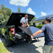 On a sunny day, two men carefully load a Solarever 450w Solar Panel from Solarever USA into the back of a black truck. One is inside the truck bed, while the other stands outside, both guiding the panel. In the background, a house with bifacial solar panels on the roof glistens in the sun.