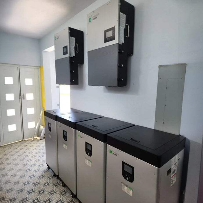 A row of four Fortress Power eVault Max 18.5kWh LFP Battery units with black lids is installed on a patterned floor. Two large inverters are mounted on the wall above. The room features light gray walls and benefits from natural light from a nearby window, ideal for solar storage with Lithium Iron Phosphate technology.